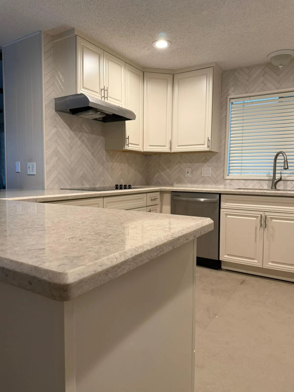 Speckled quartz peninsula with black cooktop, stainless hood, and herringbone backsplash